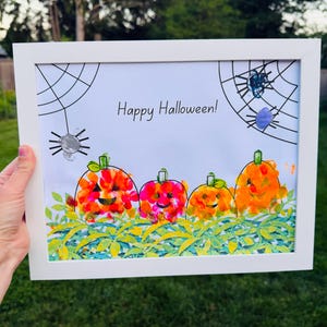 May include: A white framed print featuring the text Happy Halloween in black font. The artwork displays four colorful pumpkins with smiling faces, green leaves at the base, and black line-drawn spider webs with spiders in the upper corners.