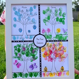 Può includere: Un'opera d'arte incorniciata intitolata "Le Quattro Stagioni" presenta quattro illustrazioni di alberi, ognuna rappresentante una stagione. Gli alberi sono decorati con impronte digitali in vari colori, con fiocchi di neve, fiori, farfalle e zucche. La cornice è bianca.
