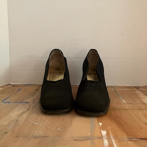 May include: A pair of black, low-heeled shoes with a rounded toe and a fabric upper. The shoes are displayed on a wooden surface. The shoes appear to be vintage or pre-owned.