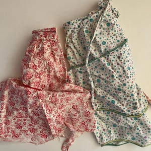 May include: Two aprons. One is white with a red floral pattern. The other is white with a pattern of teal, pink, and purple polka dots. Both aprons have ties and are made of fabric.