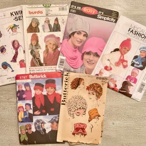 May include: A collection of six sewing pattern envelopes for hats and scarves, including brands Kwik Sew, Burda, Simplicity, McCall's, and Butterick. The envelopes feature illustrations and photographs of various headwear styles, including beanies, berets, and winter caps, with printed text identifying the pattern numbers and brand names.