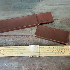 Puede incluir: Una regla de cálculo vintage con un cursor de plástico transparente, sobre una superficie de madera. Dos estuches rectangulares marrones están abiertos, uno con la etiqueta "Natacha". La regla es de color crema con marcas negras.