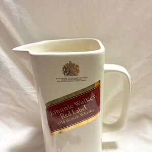 May include: Cream-colored ceramic pitcher with a handle and a pouring spout. Features the "Johnnie Walker Red Label Old Scotch Whisky" logo in red and gold. Includes the Johnnie Walker logo and "ESTABLISHED 1820" at the bottom.