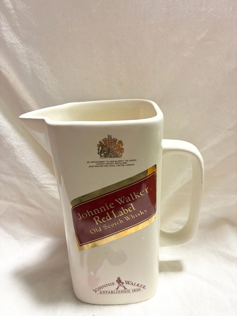 May include: Cream-colored ceramic pitcher with a handle and a pouring spout. Features the "Johnnie Walker Red Label Old Scotch Whisky" logo in red and gold. Includes the Johnnie Walker logo and "ESTABLISHED 1820" at the bottom.