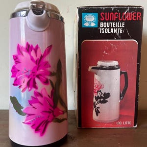 May include: A light pink insulated carafe featuring a vibrant magenta floral design and a white lid. Beside it stands its original cardboard box, which displays the text SUNFLOWER BOUTEILLE ISOLANTE and 1.00 LITRE, along with a picture of the carafe.