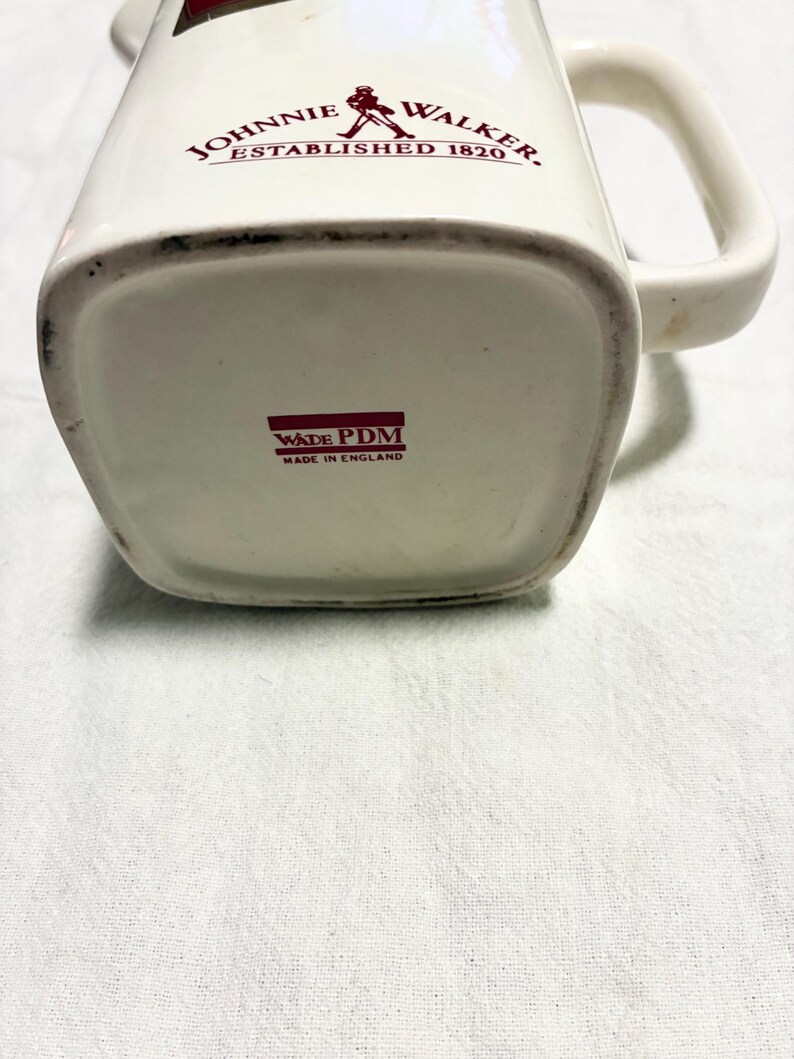 May include: Cream-colored ceramic pitcher with a handle, featuring the text "JOHNNIE WALKER ESTABLISHED 1820" in red. The bottom of the pitcher has the text "WADE PDM MADE IN ENGLAND".