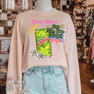 May include: A light pink sweatshirt featuring a graphic of a green Mountain Dew can with the text "Guest Check" and "One Mtn Dew Please!" in a playful font. The design includes stars and hearts.