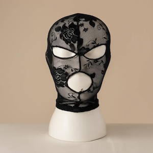 Peut inclure: Balaclava en dentelle noire avec motifs floraux, avec ouvertures pour les yeux et la bouche. Le passe-montagne est présenté sur une tête de mannequin blanche sur fond neutre. Le motif en dentelle ajoute une texture délicate.