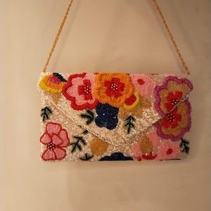 Op de afbeelding: Een met kralen bezette clutch met een gouden kettingriem. De crèmekleurige tas is versierd met kleurrijke bloemen van kralen in roze, geel, oranje en blauw. Groene kralenblaadjes accentueren het bloemmotief.