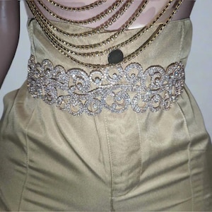 以下が含まれることがあります： ゴールドカラーのフォーマルジャンプスーツ。装飾的なラインストーンベルトと、重ね着されたゴールドチェーンネックレスが特徴です。ベルトは透明なラインストーンを使った複雑なデザインです。ジャンプスーツはストラップレスデザインです。