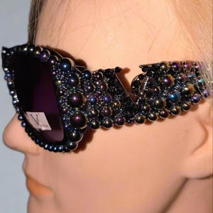 May include: Black sunglasses with dark purple lenses and a decorative frame. The frame is embellished with iridescent, dark-colored beads. The sunglasses are on a mannequin head.