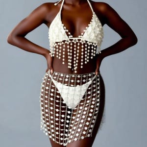 以下が含まれることがあります： アイボリーホワイトのビキニセット。ホルターネックのトップスとスカート。トップスは小さなパールと垂れ下がるストランドで装飾されています。スカートは、接続された円形の金属片で作られています。水着やビーチウェア用にデザインされています。
