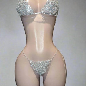 以下が含まれることがあります： きらびやかなラインストーンが施されたシルバーのビキニセット。トップはホルターネックのデザインで、ボトムはTバックです。輝く装飾が施された水着がマネキンに展示されています。