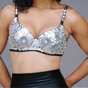 May include: A silver sequined bra with black trim and straps. The bra features a central design with larger, clear embellishments. The straps are also adorned with small, clear embellishments. The bra is worn with a black, high-waisted garment.
