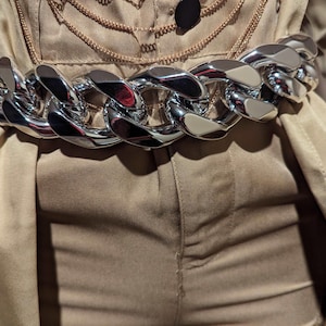 以下が含まれることがあります： 光沢のあるシルバーのチェーンベルト。大きなインターロッキングリンクが特徴です。ベージュ色の衣装の上から着用され、大胆なコントラストを生み出しています。ベルトの磨かれた表面は光を反射し、華やかさを添えています。