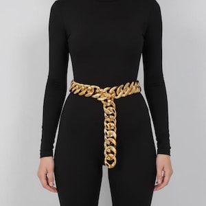 May include: A gold-colored chain belt with a large link design. The belt is worn over a black long-sleeved top and pants. The belt has a decorative chain that hangs down the front.