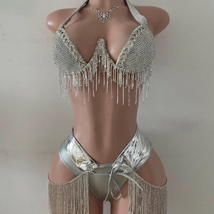 Könnte beinhalten: Silbernes zweiteiliges Outfit mit Neckholder-Top und High-Waist-Hose. Das Oberteil ist mit Strasssteinen und hängenden Fransen verziert. Die Hose hat einen metallischen Glanz und hängende Fransen an den Seiten. Ein passendes Strass-Bruststück ist ebenfalls enthalten.