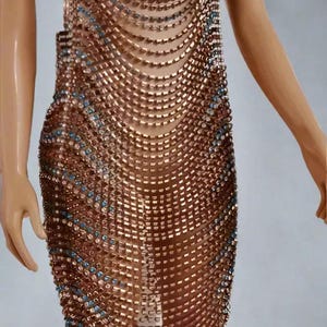 以下が含まれることがあります： 銅色のチェーンメイルドレス、ホルターネック。 小さな正方形の金属リンクで作られたドレスで、青と透明なクリスタルがアクセントになっています。 マネキンに飾られたドレスです。