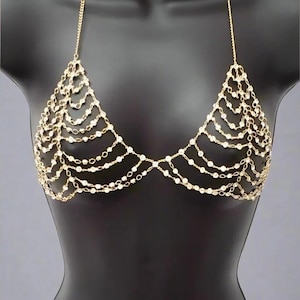 以下が含まれることがあります： ゴールドカラーのボディチェーンブラ。ホルターネックのデザインで、小さな丸い透明なビーズと繊細なチェーンリンクが複数連なり、レイヤード感を演出。黒いマネキンに展示されています。