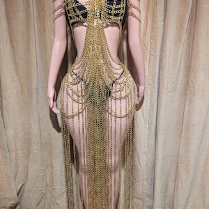 以下が含まれることがあります： ホルターネックと複雑なチェーンのディテールが特徴のゴールドチェーンのドレス。 ドレスは、流れるようなエレガントなシルエットを作り出す一連の滝のチェーンを備えています。 ドレスはマネキンに展示されています。