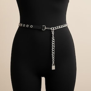以下が含まれることがあります： シルバーカラーのチェーンとバックルのディテールが施された黒いベルト。ベルトには一連のアイレットと、小さな南京錠チャームが付いたぶら下がっているチェーンが付いています。ベルトはウエストの周りに着用されています。