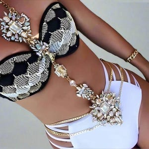 以下が含まれることがあります： 黒と白の模様のビキニトップ、ゴールドチェーンと宝石の装飾付き。ビキニボトムは白で、ゴールドのストラップが付いています。腕にはゴールドのブレスレットが見えます。