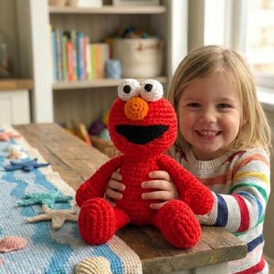 Könnte beinhalten: Eine handgefertigte, rote Elmo-Puppe aus Häkelarbeit mit weißen Augen und einer orangefarbenen Nase. Das Kind hält das Spielzeug lächelnd. Im Hintergrund ein Holztisch, ein Regal mit Büchern und ein blau-weiß gehäkelter Tischläufer mit Muschelakzenten.
