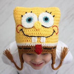 May include: A yellow, white, and brown crocheted hat designed to resemble Spongebob Squarepants. The hat features large white eyes with blue irises, a wide black stitched smile, two white teeth, and a red tie detail, with braided earflaps.
