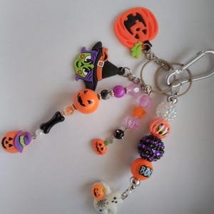 Puede incluir: Un llavero temático de Halloween con varios dijes. Incluye calabazas naranjas, un sombrero de bruja, un fantasma y cuentas en naranja, morado y rosa. La palabra "BOO" está en uno de los dijes de calabaza. El llavero tiene un cierre plateado.