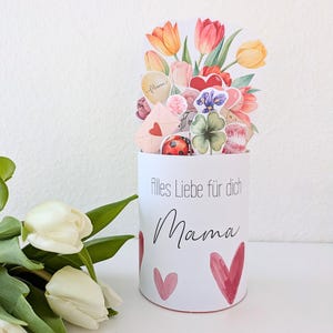 Könnte beinhalten: Eine weiße zylindrische Karte mit den Worten "Alles Liebe für dich Mama" und roten Herzen. Die Karte ist mit Papierschnitten von Tulpen, Herzen, einem Marienkäfer und einem vierblättrigen Kleeblatt verziert. Ein Strauß weißer Tulpen befindet sich im Vordergrund.