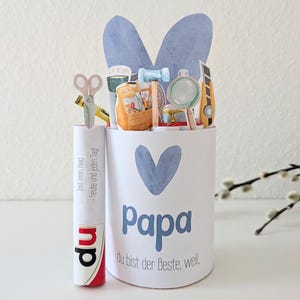 May include: A white cylindrical container with a blue heart and the word "Papa" in blue. The container is decorated with paper cutouts of tools and a pair of blue bunny ears. A tube of glue is next to the container with text.
