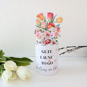 Puede incluir: Un recipiente cilíndrico blanco con un arreglo floral emergente con tulipanes, margaritas y rosas. El recipiente tiene el texto "GUTE LAUNE TO GO" y una ilustración de mariquita. Lunares rosas decoran el recipiente.