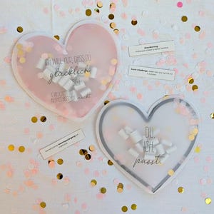Puede incluir: Dos decoraciones en forma de corazón con texto y confeti. Una es rosa y la otra blanca. El corazón rosa dice "ICH WILL NUR, DASS DU glücklich BIST & VIELLEICHT EIN KLEINES BISSCHEN nackt." El corazón blanco dice "DU. ICH. passt."