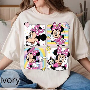 以下が含まれることがあります： アイボリー色のTシャツ。4つのパネルにミニーマウスのイラストが描かれています。各パネルには、ピンクのドット柄のリボンとドレスを着たミニーが様々なポーズで描かれています。背景パネルには、ハート、虹、星など、カラフルなデザインが施されています。