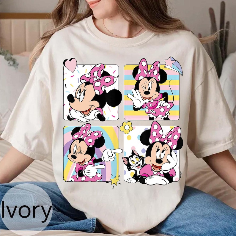 以下が含まれることがあります： アイボリー色のTシャツ。4つのパネルにミニーマウスのイラストが描かれています。各パネルには、ピンクのドット柄のリボンとドレスを着たミニーが様々なポーズで描かれています。背景パネルには、ハート、虹、星など、カラフルなデザインが施されています。
