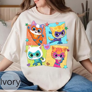 May include: Ivory-colored t-shirt featuring a colorful graphic of four superhero cats in square frames. Each cat is in a different pose and wearing a mask and costume. The background of each square is a different color, with stars and hearts.