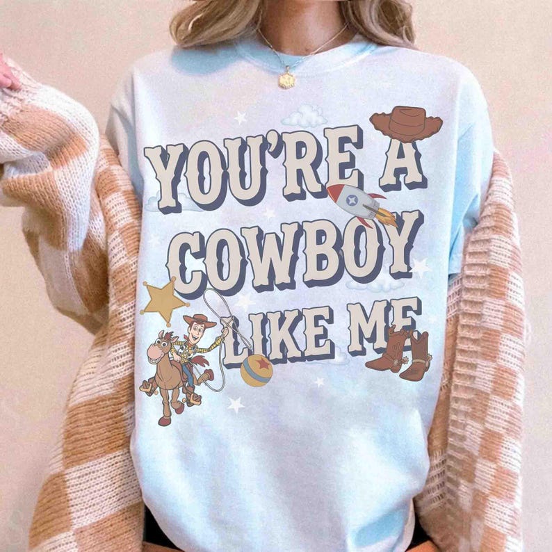 May include: A light blue t-shirt featuring the text YOU'RE A COWBOY LIKE ME in a bold font. The design includes a cartoon cowboy on a horse, a brown hat, a rocket, a yellow star, a ball, and brown boots scattered across the front of the shirt.