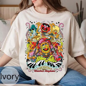 Könnte beinhalten: Elfenbeinfarbenes T-Shirt mit einer farbenfrohen Grafik der Muppets-Band Dr. Teeth and The Electric Mayhem. Das Design zeigt die Bandmitglieder beim Spielen verschiedener Instrumente, mit dem Bandnamen in einer stilisierten Schriftart.