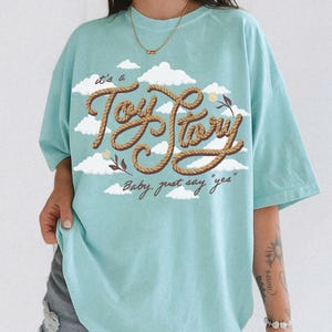 May include: A light blue t-shirt featuring a graphic print of white clouds and the text "it's a Toy Story" written in a brown rope-style font, with the additional text "Baby, just say "yes"" printed below in a smaller brown cursive script.