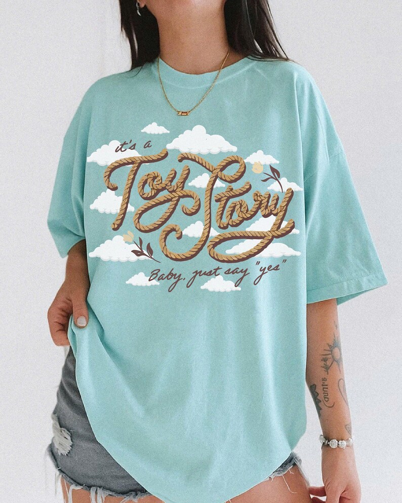May include: A light blue t-shirt featuring a graphic print of white clouds and the text "it's a Toy Story" written in a brown rope-style font, with the additional text "Baby, just say "yes"" printed below in a smaller brown cursive script.