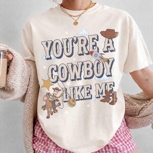 May include: A cream-colored t-shirt featuring the text "YOU'RE A COWBOY LIKE ME" in a bold, dark blue font. The graphic includes a cartoon cowboy on a horse, a rocket ship, a brown hat, a yellow star, a ball, and small white clouds and stars.