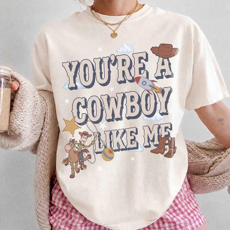 May include: A cream-colored t-shirt featuring the text "YOU'RE A COWBOY LIKE ME" in a bold, dark blue font. The graphic includes a cartoon cowboy on a horse, a rocket ship, a brown hat, a yellow star, a ball, and small white clouds and stars.