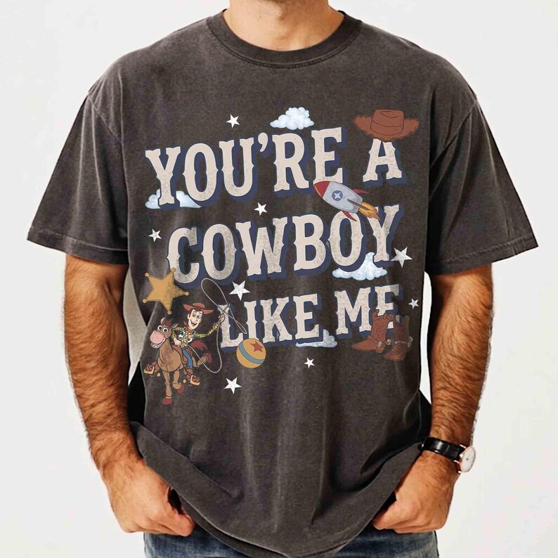 May include: A charcoal gray short-sleeve t-shirt featuring the text "YOU'RE A COWBOY LIKE ME" in a distressed white font. The graphic includes a cartoon cowboy on a horse, a rocket, a brown hat, a star, a ball, and small white stars and clouds.