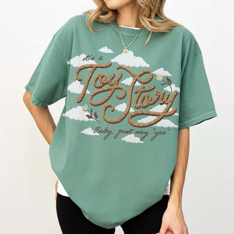 May include: A sage green short-sleeve t-shirt featuring a graphic print of white clouds and brown rope-textured lettering that reads "it's a Toy Story" and "Baby, just say "yes"" with small floral accents on the front of the garment.
