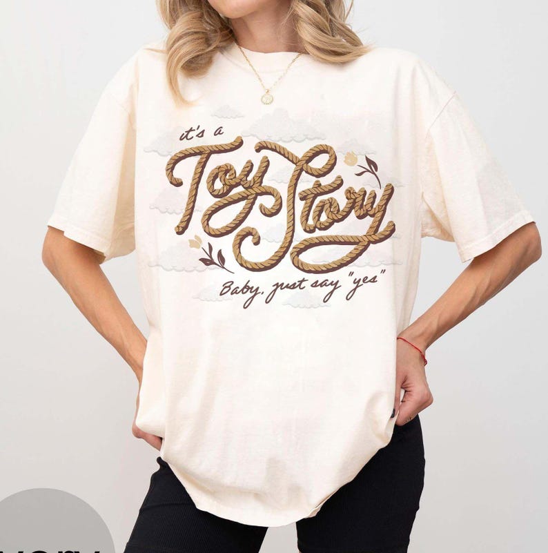 May include: A cream-colored short-sleeve t-shirt featuring a graphic print of the text "it's a Toy Story" in a brown rope-like font, accompanied by the phrase "Baby, just say "yes"" and small illustrations of clouds and flowers.