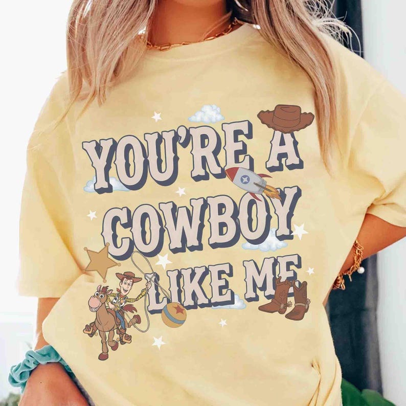 May include: A pale yellow t-shirt featuring the text "YOU'RE A COWBOY LIKE ME" in a bold, dark font. The graphic includes a cartoon cowboy riding a horse, a rocket ship, a brown hat, a pair of brown boots, a star, a ball, and small white stars.