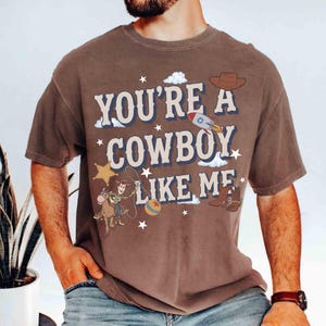 May include: A brown short-sleeve t-shirt featuring the text "YOU'RE A COWBOY LIKE ME" in large cream letters. The graphic includes a cartoon cowboy on a horse, a rocket, a star, a hat, and boots, surrounded by small white stars and clouds.
