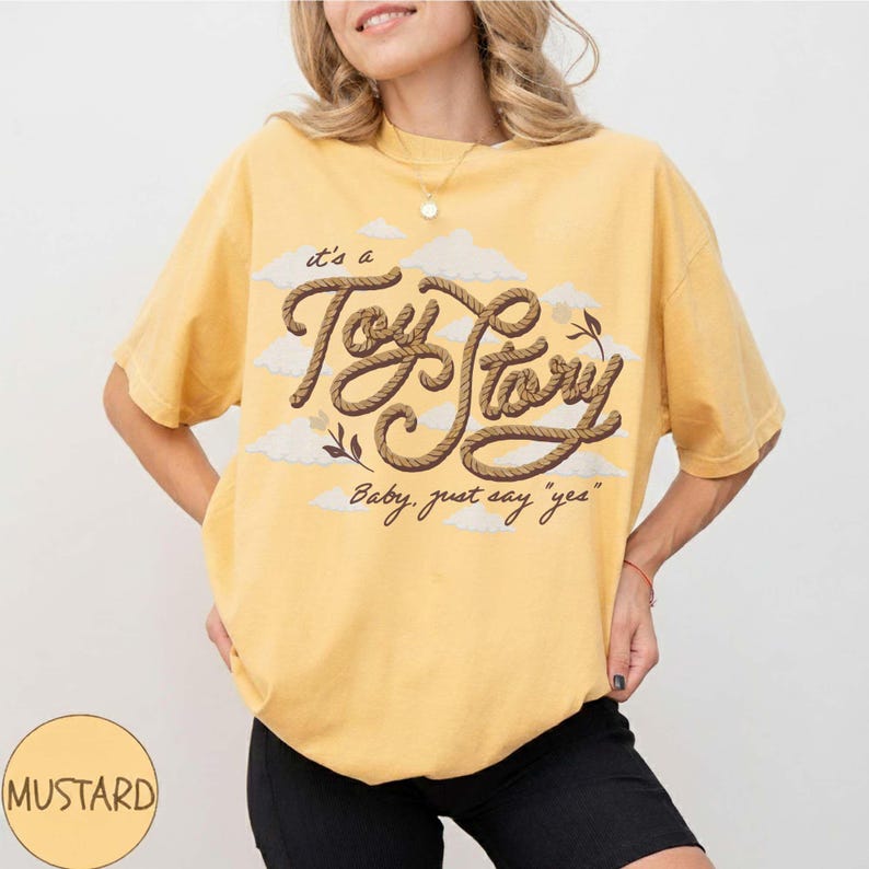 May include: A mustard yellow short-sleeve t-shirt featuring a graphic of brown rope lettering that reads "it's a Toy Story" and "Baby, just say "yes"" with white cloud illustrations in the background. The shirt has a crew neckline and a loose fit.