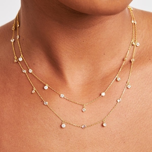 Könnte beinhalten: Zwei zarte goldene Halsketten mit kleinen, klaren Edelsteinen. Die Halsketten sind geschichtet, wobei eine etwas länger ist als die andere. Die Edelsteine sind gleichmäßig entlang der Ketten angeordnet. Schmuck.