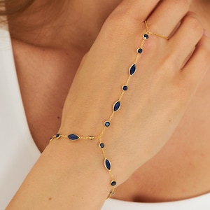 May include: A gold-coloured hand chain bracelet with a delicate design. The bracelet features a series of dark blue, oval and round gemstones connected by a thin chain. The bracelet extends from the wrist to the finger.